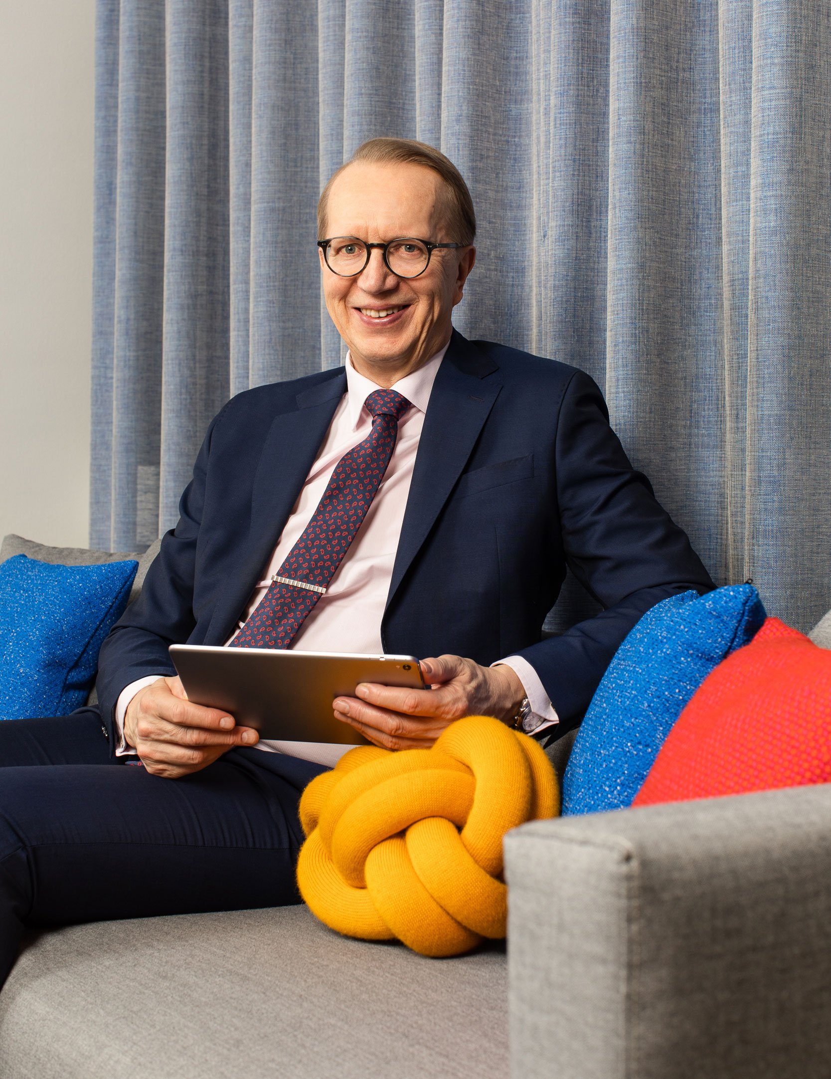 Työllisyysrahaston toimitusjohtaja Janne Metsämäki istuu sohvalla katsoo kameraan ja hymyilee. Hänelle on tumma puku päällään ja älylaite kädessään.